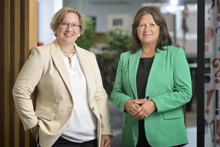 Silvia Hruška-Frank und Renate Anderl stehen in einem modernen Büro und blicken freundlich in die Kamera. Silvia Hruška-Frank trägt eine Brille, ein weißes Oberteil und einen cremefarbenen Blazer, während Renate Anderl einen grünen Blazer mit schwarzem Oberteil trägt. Eine lehnt an einer Holzsäule, die andere hat die Hände gefaltet. Im Hintergrund sind Pflanzen und Büroeinrichtungen sichtbar.