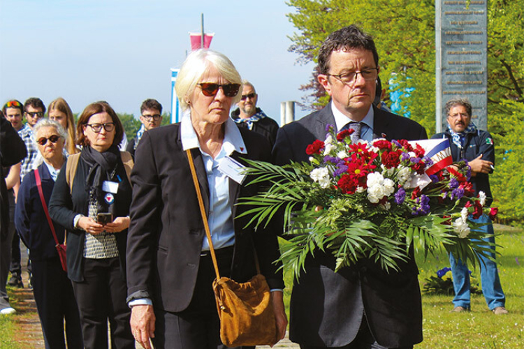 People dressed in black are attending a memorial event; two individuals in the foreground are jointly carrying a floral arrangement with red, white, and purple flowers.