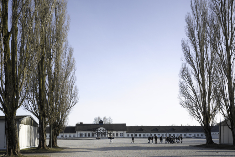 Gedenkstätte des ehemaligen Konzentrationslagers Dachau: Blick auf den Appellplatz mit kahlen Pappeln und historischen Baracken bei klarem Himmel. Eine Besuchergruppe geht auf das zentrale Museumsgebäude zu. © Stefan Müller-Naumann/KZ-Gedenkstätte Dachau