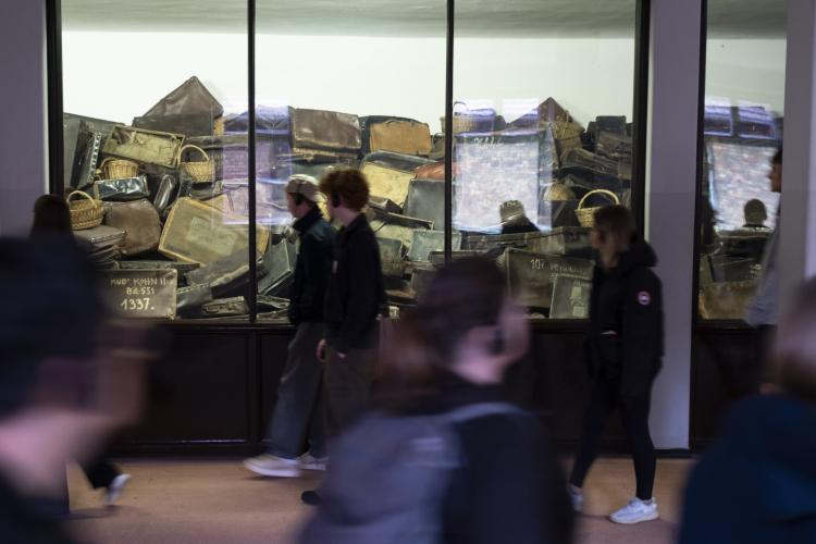 Besucher:innen gehen an einer Vitrine im Auschwitz-Museum vorbei, in der eine große Anzahl alter Koffer und Körbe gestapelt ist – persönliche Gegenstände von Holocaust-Opfern, die an deren Ankunft im Konzentrationslager erinnern. Die Szene vermittelt einen stillen, nachdenklichen Moment im Gedenken an die Geschichte.