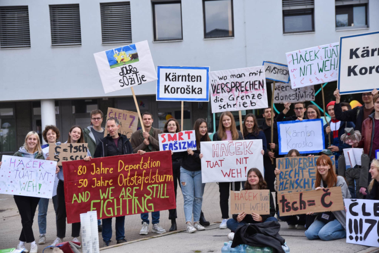 Eine Gruppe junger Menschen steht vor einem modernen Gebäude und hält Protestschilder hoch. Die Plakate sind zweisprachig (Deutsch und Slowenisch) und thematisieren Minderheitenrechte, den sogenannten „Ortstafelstreit“ in Kärnten sowie die Bedeutung slowenischer Sonderzeichen wie dem Háček. Die Stimmung ist friedlich und engagiert. © Franc Wakounig