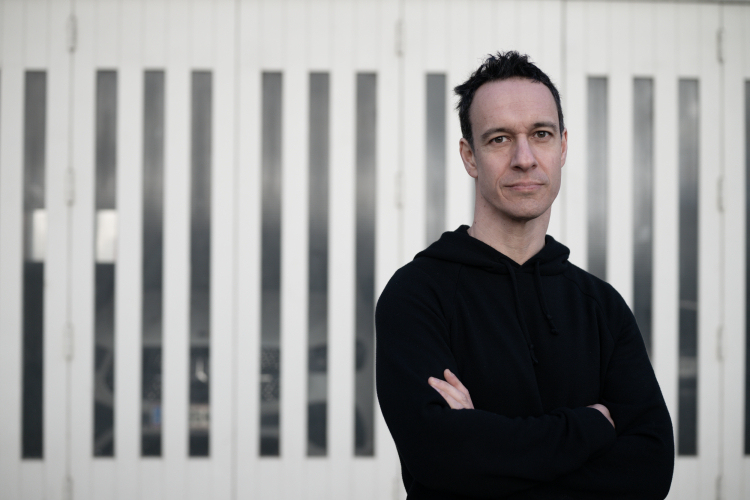 Bernhard Weidinger (kurze dunkle Haare) trägt einen schwarzen Hoodie und steht mit verschränkten Armen vor einer weißen Wand mit vertikalen Glaselementen.